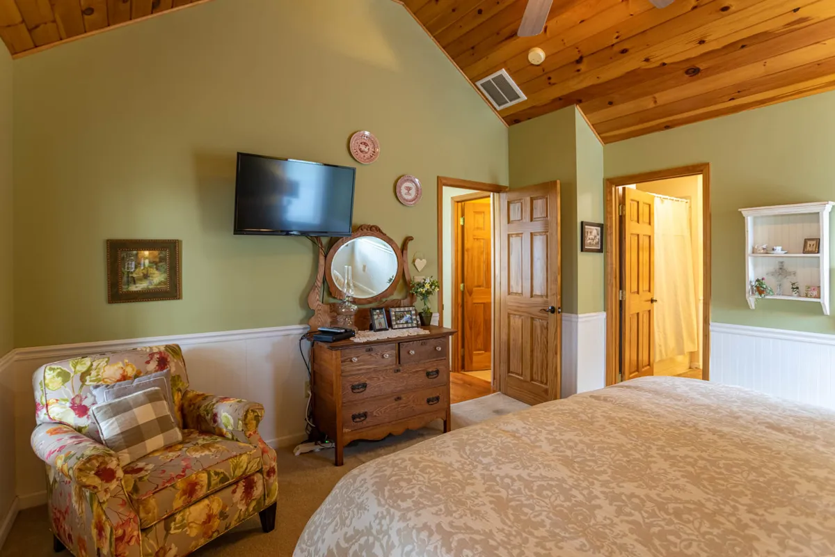 A cozy corner with a floral armchair, a wooden dresser, a wall-mounted TV, and decorative plates creates an intimate setting for morning coffee or quiet TV evenings. Sunlight streams into the space, enhancing its inviting atmosphere.
