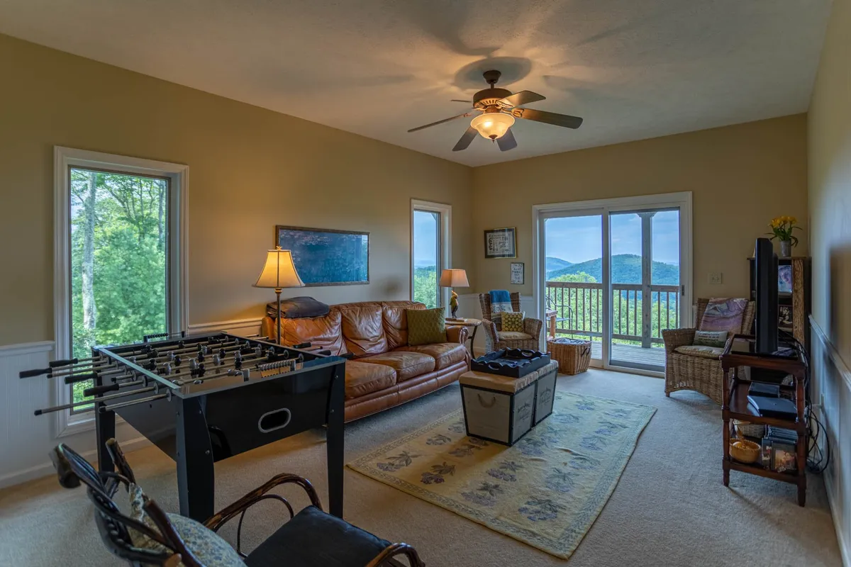 This image showcases a spacious lower-level living area featuring a foosball table, large windows with scenic views, a comfortable leather sofa, a coffee table, and a wall-mounted TV. The scene is casual and social, reflecting a fun, laid-back atmosphere ideal for gatherings or game nights.