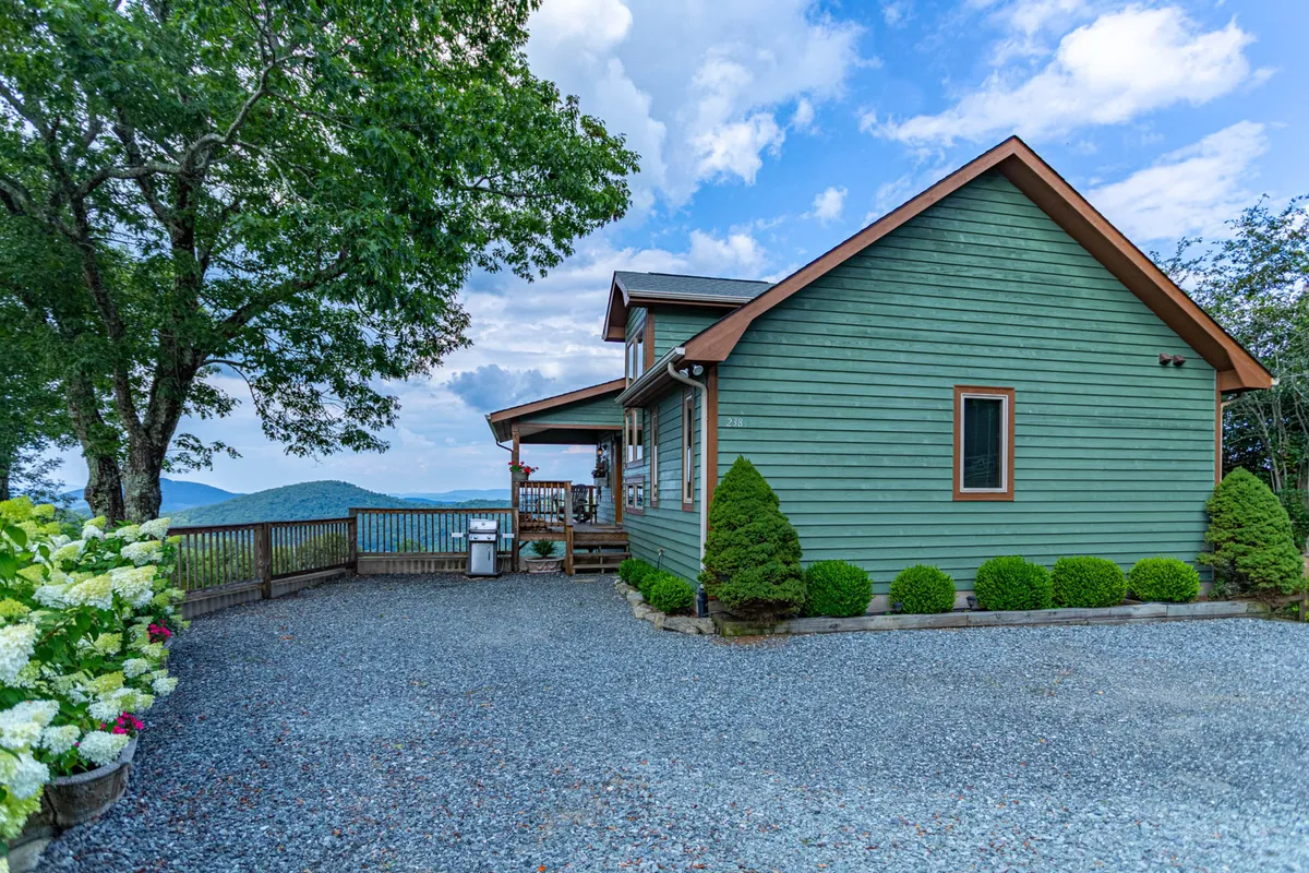 This image shows the exterior of the green house surrounded by trees and flower beds, with a gravel driveway and a backdrop of mountains. The deck and outdoor space are visible, suggesting areas for group gatherings or children to play.