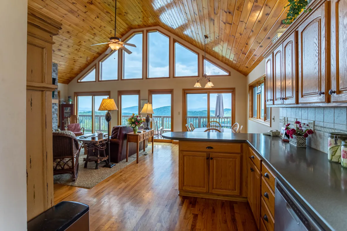 A cozy interior living space with large windows framing sweeping mountain views. The room features a wooden ceiling, a kitchen island, and a comfortable seating area—inviting guests to relax and enjoy the breathtaking scenery, especially as the sun sets.