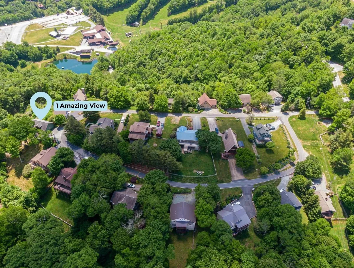 A tranquil exterior scene shows homes nestled among trees with a winding road, a peaceful pond, and a dramatic backdrop of mountain ridges. The setting sun casts a warm glow across the landscape, highlighting the secluded, natural beauty of the area.