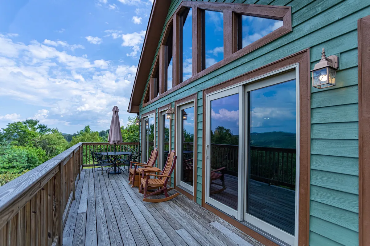 A wide, sunlit view of the cabin's large wooden deck, complete with an outdoor dining table, rocking chairs, and an expansive mountain vista beyond. The deck wraps around the house, blending seamlessly with floor-to-ceiling glass windows that invite the outdoors in. The setting is tranquil, perfect for meals, relaxing, or simply admiring the shifting mountain scenery.