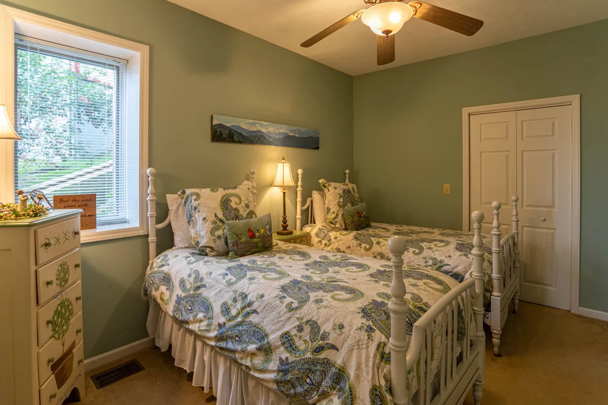 A welcoming bedroom with two twin beds adorned with decorative bedding, a lamp on the nightstand, natural light streaming through a window, and wall art depicting mountain scenery. The scene is both cheerful and soothing, inviting family or friends to relax and share stories.