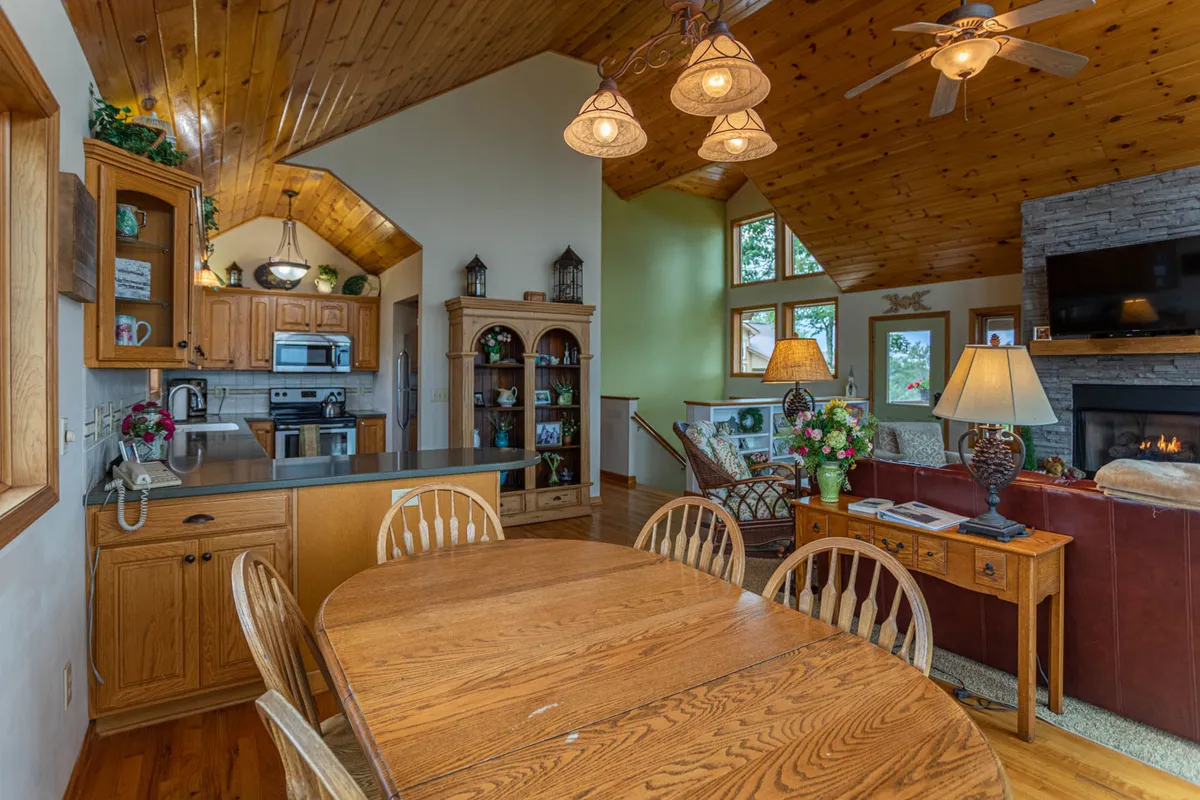 A panoramic shot encompassing the dining table, kitchen cabinetry, plush living room seating, and the fireplace, all set against a backdrop of large windows that reveal scenic mountain vistas. The warm and inviting space is ready for family gatherings, cooking, and relaxing by the fire.