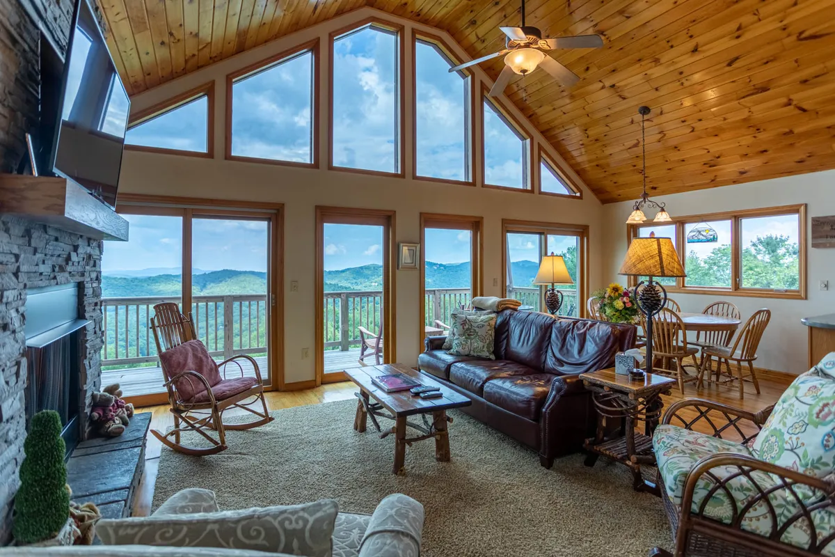 A spacious open-concept living and dining area featuring comfortable leather sofas, a striking stone fireplace, a high wooden ceiling, and expansive windows that flood the space with natural light and provide breathtaking mountain views. The dining table is perfectly positioned to enjoy the scenery, and the overall setup is ideal for gatherings and cozy evenings.
