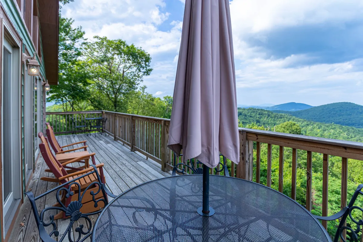 A picturesque setting on the deck with an outdoor table shaded by an umbrella, surrounded by wooden rocking chairs. The table is perfectly positioned for al fresco dining against a backdrop of rolling mountains and open sky, inviting guests to gather for meals or sunset conversations.