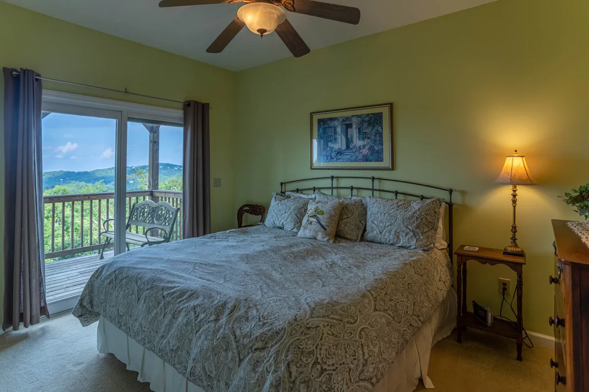 A tranquil bedroom scene with a large bed, decorative lamp, captivating artwork, and expansive windows offering calming views. Natural light filters into the room, enhancing its soothing atmosphere—perfect for a restful night or a peaceful morning routine.