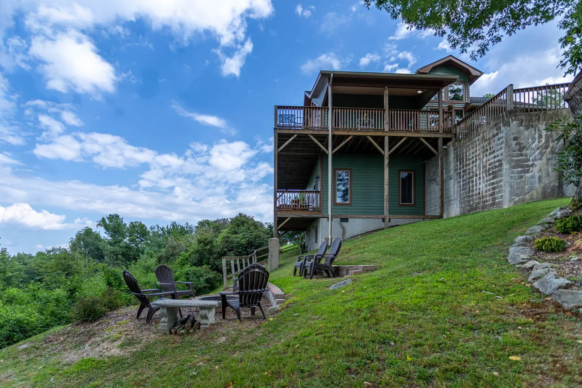 An exterior view showing the house structure, spacious deck area, a cozy fire pit, surrounding trees, and a sweeping lawn. The scene suggests opportunities for outdoor gatherings, barbecues, and relaxing by the fire while enjoying nature and mountain views.