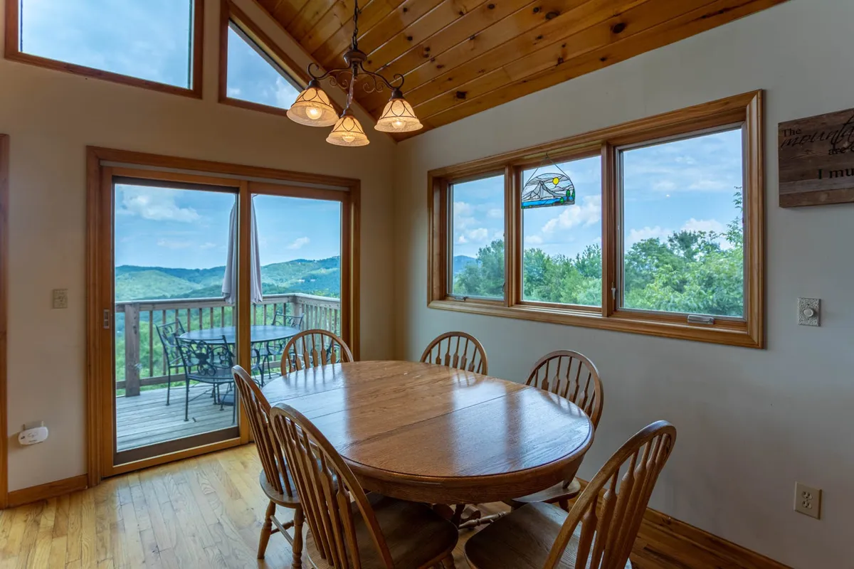 This image shows a warm, inviting dining area with a large wooden table, plenty of natural light streaming through expansive windows, and a clear view of the outdoor deck. The stained glass window adds character, and the connection between the dining space and the adjacent deck is visually apparent.